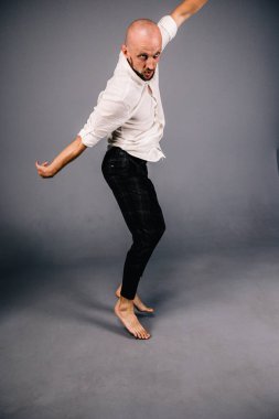 Beyaz gömlekli ve ekose pantolonlu, gri arka planda dans eden güzel sakallı, kel bir adamın stüdyo portresi.