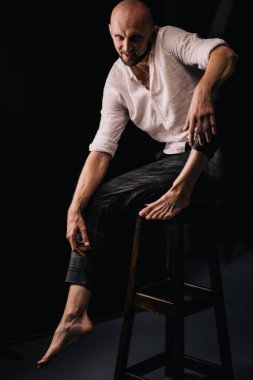 Beyaz gömlekli ve ekose pantolonlu, siyah arka planda bar taburesinde oturan kel bir adam.