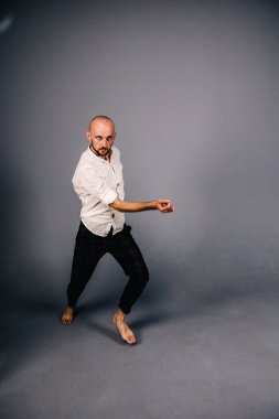 Beyaz gömlekli ve ekose pantolonlu, gri arka planda dans eden güzel sakallı, kel bir adamın stüdyo portresi.