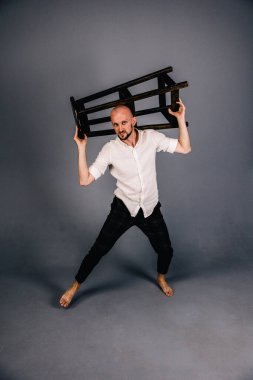 Çıplak ayaklı, kel, sakallı, beyaz gömlekli, ekose pantolonlu, gri arka planda bar taburesiyle poz veren bir adam..