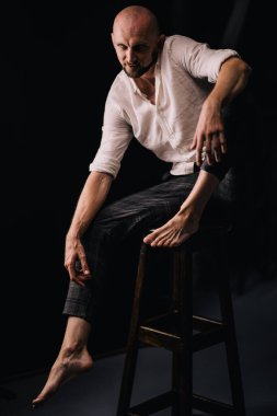 Beyaz gömlekli ve ekose pantolonlu, siyah arka planda bar taburesinde oturan kel bir adam.