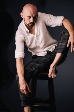Beyaz gömlekli ve ekose pantolonlu, siyah arka planda bar taburesinde oturan kel bir adam.