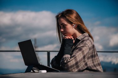 Ekose gömlekli, genç, ince bir kız doğada bir dizüstü bilgisayarda, dağların arka planına karşı çalışıyor.