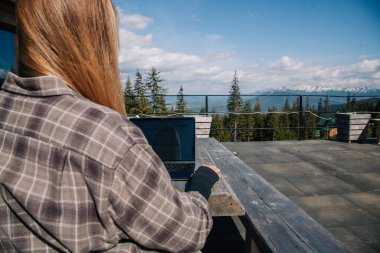 Ekose gömlekli genç, kızıl saçlı bir kız, dağların arka planına karşı eski bir ahşap masada oturur ve bir dizüstü bilgisayar üzerinde çalışır. Daktilo. Arkadan görünüm.