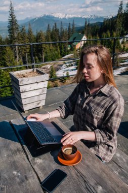 Uzun saçlı, genç, güzel bir kız açık havada eski bir ahşap masada oturur, kahve içer ve açık havada dizüstü bilgisayarının önünde dağların arka planına karşı çalışır. Üst görünüm