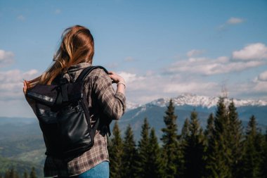 A young, slender girl with loose hair in a plaid shirt and jeans with a black backpack stands against the backdrop of mountains in sunny weather. View from the back.