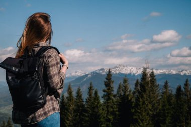 A young, slender girl with loose hair in a plaid shirt and jeans with a black backpack stands against the backdrop of mountains in sunny weather. View from the back.