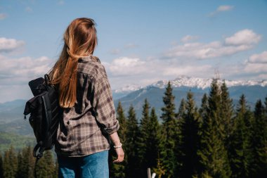 A young, slender girl with loose hair in a plaid shirt and jeans with a black backpack stands against the backdrop of mountains in sunny weather. View from the back.