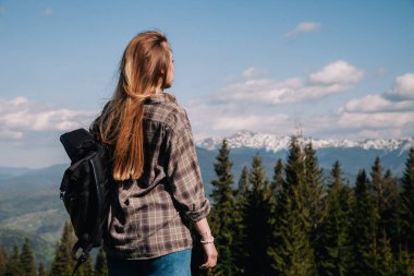 A young, slender girl with loose hair in a plaid shirt and jeans with a black backpack stands against the backdrop of mountains in sunny weather. View from the back.