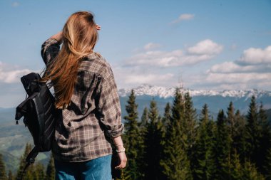 A young, slender girl with loose hair in a plaid shirt and jeans with a black backpack stands against the backdrop of mountains in sunny weather. View from the back.