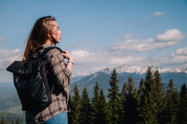 A young, slender girl with loose hair in a plaid shirt and jeans with a black backpack stands against the backdrop of mountains in sunny weather. View from the back.