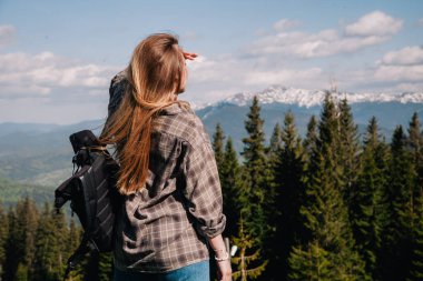 A young, slender girl with loose hair in a plaid shirt and jeans with a black backpack stands against the backdrop of mountains in sunny weather. View from the back.
