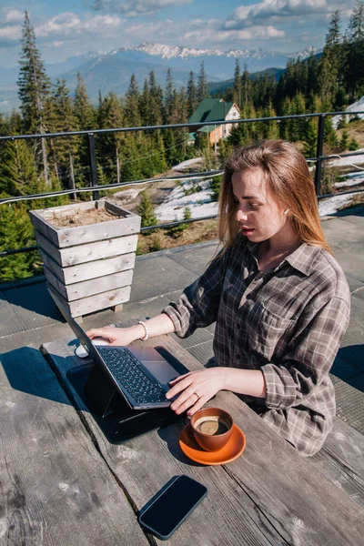 A young, beautiful girl with long hair sits at an old wooden table in the open air, drinking coffee and working at her laptop against the backdrop of the mountains in clear weather. Top view — Φωτογραφία Αρχείου