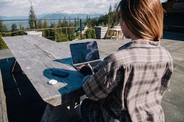 A young, beautiful girl in a plaid shirt works on a laptop at a wooden old table in nature against the backdrop of mountains, next to a smartphone and headphones. View from the back. — Φωτογραφία Αρχείου