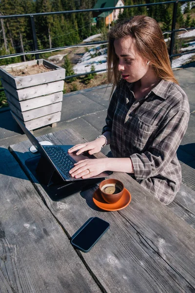 A young, beautiful girl with long hair sits at an old wooden table in the open air, drinking coffee and working at her laptop against the backdrop of the mountains in clear weather. Top view — Φωτογραφία Αρχείου