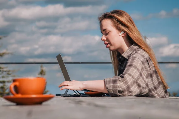 Ένα νεαρό, όμορφο κορίτσι με καρό πουκάμισο μιλάει σε ένα smartphone κάθεται σε ένα παλιό ξύλινο τραπέζι μπροστά από ένα φορητό υπολογιστή στη φύση και πίνει καφέ — Φωτογραφία Αρχείου