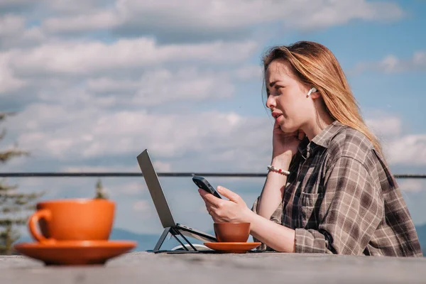 A young, beautiful girl in a plaid shirt talks on a smartphone sitting at an old wooden table in front of a laptop in nature and drinks coffee — Zdjęcie stockowe