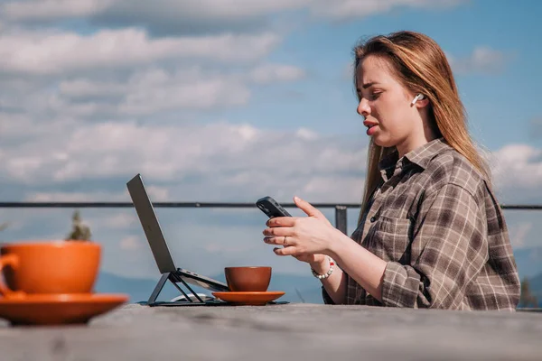 A young, beautiful girl in a plaid shirt talks on a smartphone sitting at an old wooden table in front of a laptop in nature and drinks coffee — Zdjęcie stockowe