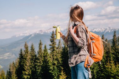 Ekose gömlekli, bol saçlı, turuncu sırt çantalı genç bir kız Karpatlar 'daki dağların arka planında parlak bir termal bardaktan çay içiyor. Arkadan görünüm.