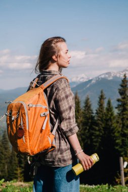 Ekose gömlekli, bol saçlı, turuncu sırt çantalı genç bir kız Karpatlar 'daki dağların arka planında parlak bir termal bardaktan çay içiyor. Arkadan görünüm.
