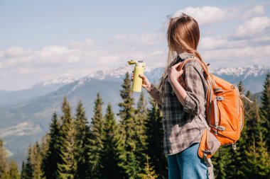 A young, girl in a plaid shirt and jeans with loose hair with an orange backpack drinks tea from a bright thermal cup against the backdrop of the mountains in the Carpathians. view from the back.