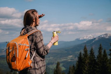 A young, girl in a plaid shirt and jeans with loose hair with an orange backpack drinks tea from a bright thermal cup against the backdrop of the mountains in the Carpathians. view from the back.