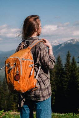Ekose gömlekli, bol saçlı, turuncu sırt çantalı genç bir kız Karpatlar 'daki dağların arka planında parlak bir termal bardaktan çay içiyor. Arkadan görünüm.