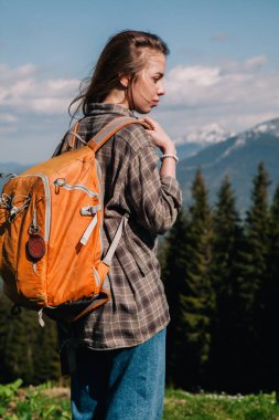 Ekose gömlekli, bol saçlı, turuncu sırt çantalı genç bir kız Karpatlar 'daki dağların arka planında parlak bir termal bardaktan çay içiyor. Arkadan görünüm.