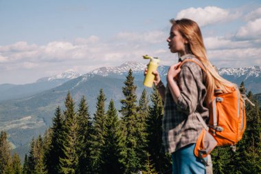 Ekose gömlekli, bol saçlı, turuncu sırt çantalı genç bir kız Karpatlar 'daki dağların arka planında parlak bir termal bardaktan çay içiyor. Arkadan görünüm.