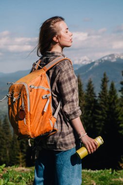 Ekose gömlekli, bol saçlı, turuncu sırt çantalı genç bir kız Karpatlar 'daki dağların arka planında parlak bir termal bardaktan çay içiyor. Arkadan görünüm.