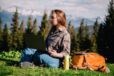 Ekose gömlek ve turuncu sırt çantalı bol saçlı genç, narin bir kız, yeşil çimlerin üzerinde, dağların arka planında oturuyor..