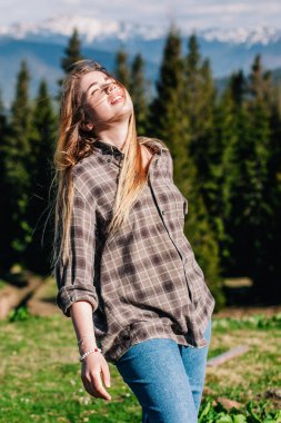 Genç, ince saçlı, ekose gömlekli ve kot pantolonlu bir kız dağlarda güneşli hava için poz veriyor.