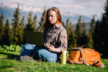 Ekose gömlek ve turuncu sırt çantalı bol saçlı genç, narin bir kız, yeşil çimlerin üzerinde, dağların arka planında oturuyor..