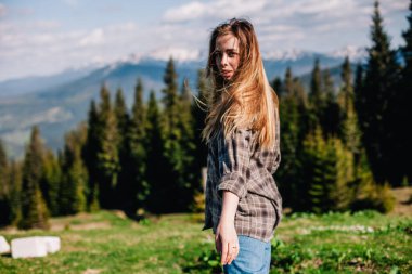 Genç, ince saçlı, ekose gömlekli ve kot pantolonlu bir kız dağlarda güneşli hava için poz veriyor.