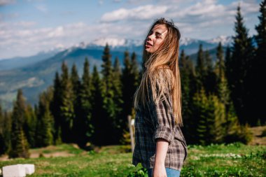 Genç, ince saçlı, ekose gömlekli ve kot pantolonlu bir kız dağlarda güneşli hava için poz veriyor.