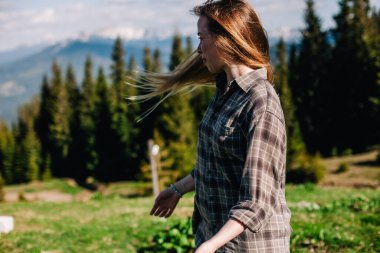 Genç, ince saçlı, ekose gömlekli ve kot pantolonlu bir kız dağlarda güneşli hava için poz veriyor.