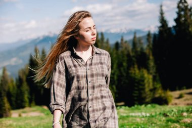 Genç, ince saçlı, ekose gömlekli ve kot pantolonlu bir kız dağlarda güneşli hava için poz veriyor.