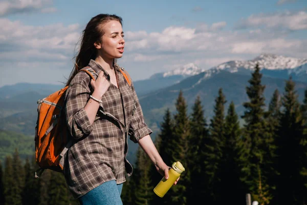Ekose gömlekli, bol saçlı, turuncu sırt çantalı genç bir kız Karpatlar 'daki dağların arka planında parlak bir termal bardaktan çay içiyor. Arkadan görünüm.