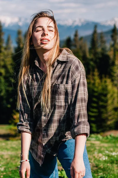 A young, slender girl with loose hair in a plaid shirt and jeans poses for sunny weather in the mountains — Photo