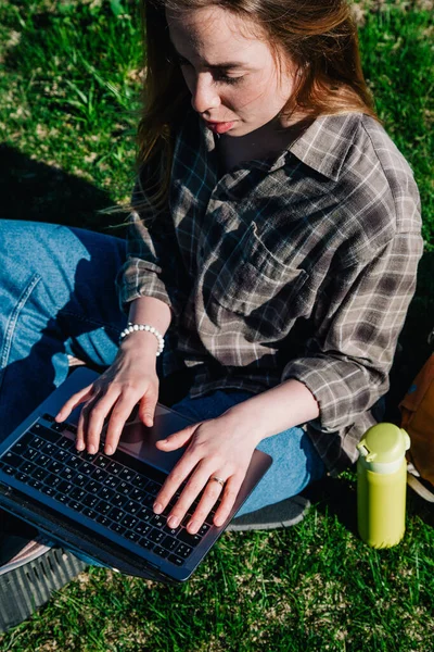 Ekose gömlek ve turuncu sırt çantalı bol saçlı genç, ince bir kız, yeşil çimlerin üzerinde, dağların arka planında oturuyor. Yukarıdan görünüm