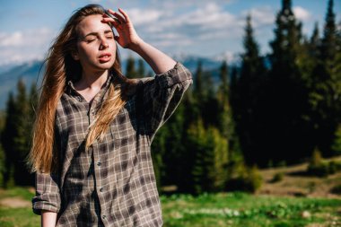 Genç, ince saçlı, ekose gömlekli ve kot pantolonlu bir kız dağlarda güneşli hava için poz veriyor.