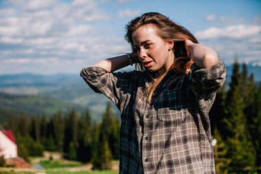 Genç, ince saçlı, ekose gömlekli ve kot pantolonlu bir kız dağlarda güneşli hava için poz veriyor.