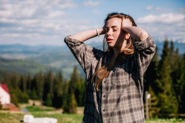 Genç, ince saçlı, ekose gömlekli ve kot pantolonlu bir kız dağlarda güneşli hava için poz veriyor.