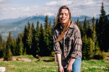 Genç, ince saçlı, ekose gömlekli ve kot pantolonlu bir kız dağlarda güneşli hava için poz veriyor.