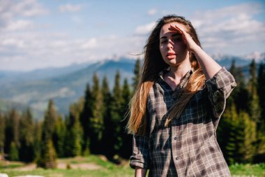 Genç, ince saçlı, ekose gömlekli ve kot pantolonlu bir kız dağlarda güneşli hava için poz veriyor.