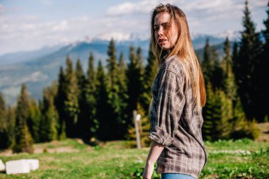 Genç, ince saçlı, ekose gömlekli ve kot pantolonlu bir kız dağlarda güneşli hava için poz veriyor.