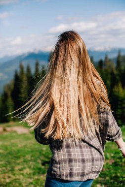 Genç, ince saçlı, ekose gömlekli ve kot pantolonlu bir kız dağlarda güneşli hava için poz veriyor.