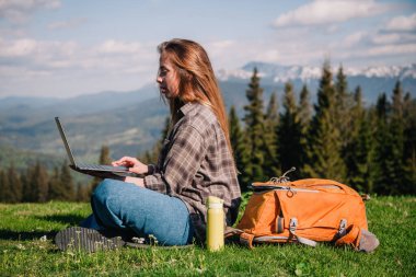 Ekose gömlek ve turuncu sırt çantalı bol saçlı genç, narin bir kız, yeşil çimlerin üzerinde, dağların arka planında oturuyor..