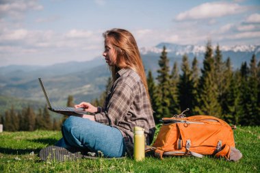 Ekose gömlek ve turuncu sırt çantalı bol saçlı genç, narin bir kız, yeşil çimlerin üzerinde, dağların arka planında oturuyor..