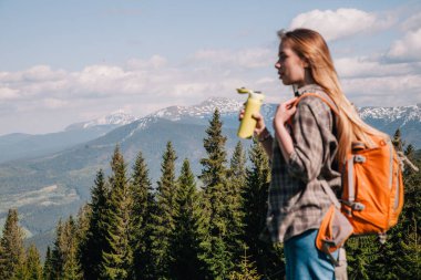 Ekose gömlekli, bol saçlı, turuncu sırt çantalı genç bir kız Karpatlar 'daki dağların arka planında parlak bir termal bardaktan çay içiyor. Arkadan görünüm.